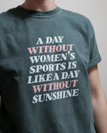 A person wearing a dark teal graphic t-shirt with the text 'A DAY WITHOUT WOMEN'S SPORTS IS LIKE A DAY WITHOUT SUNSHINE' printed in white and light pink lettering.