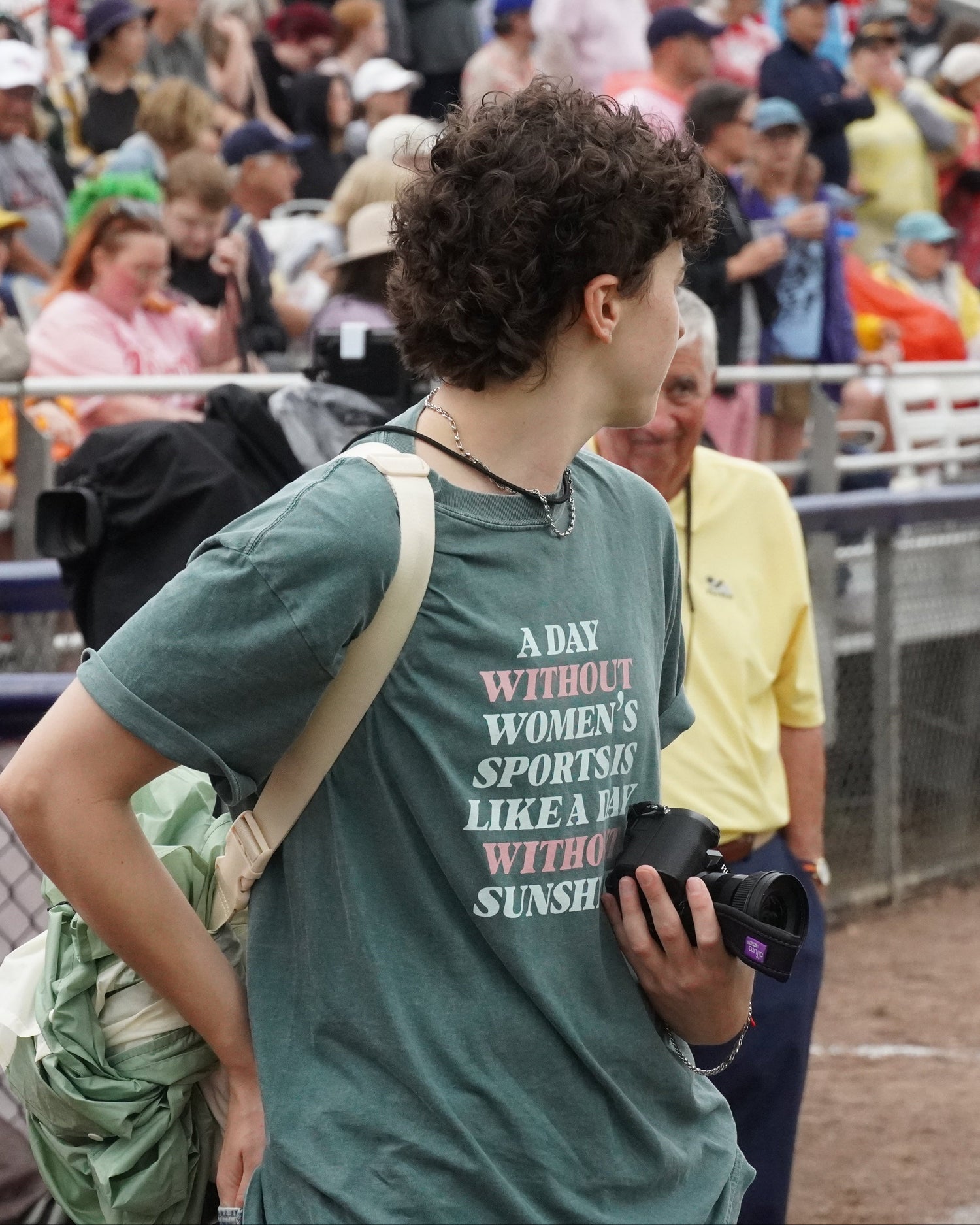 A woman at a baseball game wearing a green graphic t-shirt with the text 'A DAY WITHOUT WOMEN'S SPORTS IS LIKE A DAY WITHOUT SUNSHINE'