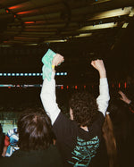 A person in a crowded indoor arena at MSG raises both arms, wearing a black T-shirt with a teal graphic and holding a teal towel overhead. Other people are visible around them, and the venue interior with lighting rigs and screens appears in the background.