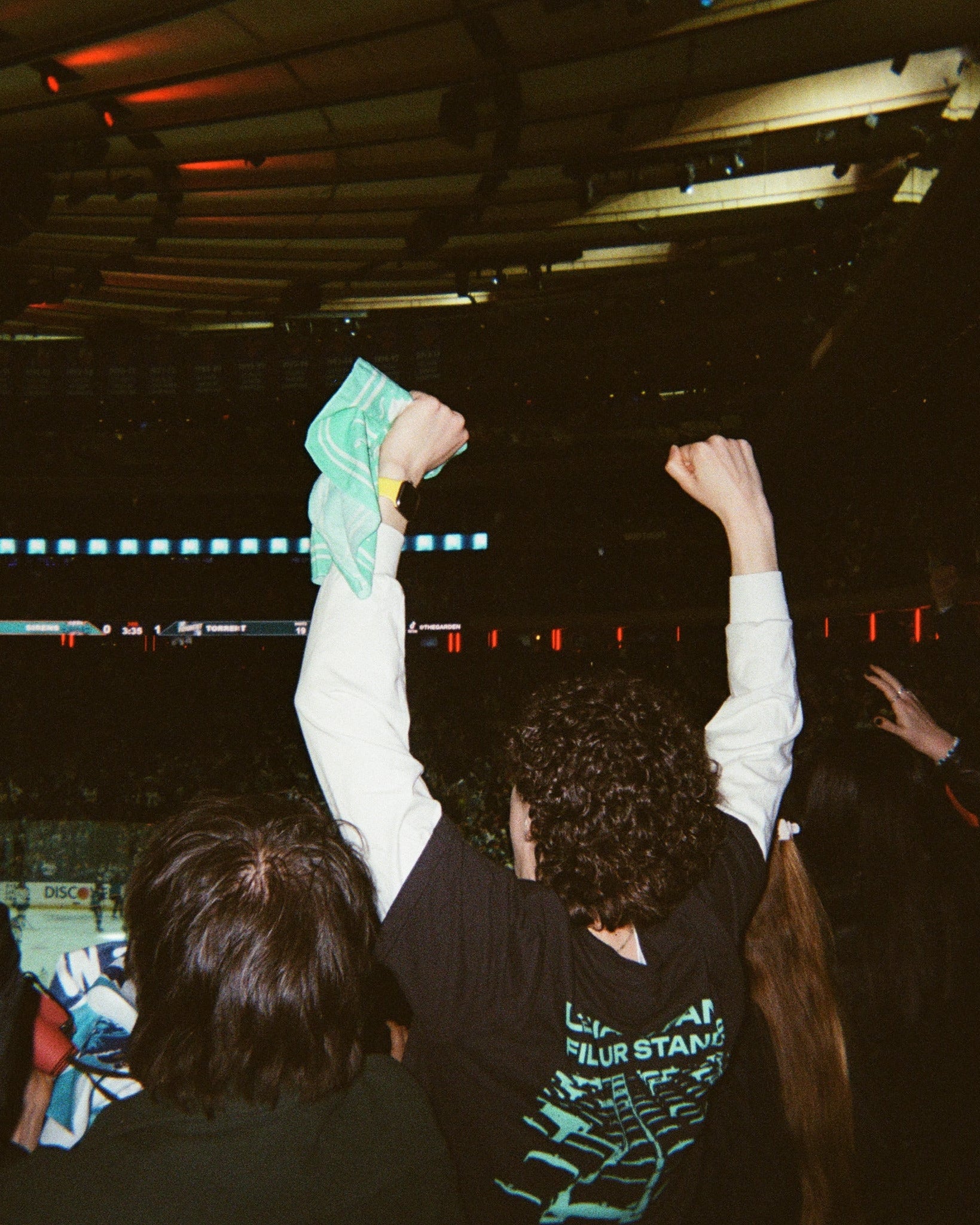 A person in a crowded indoor arena at MSG raises both arms, wearing a black T-shirt with a teal graphic and holding a teal towel overhead. Other people are visible around them, and the venue interior with lighting rigs and screens appears in the background.