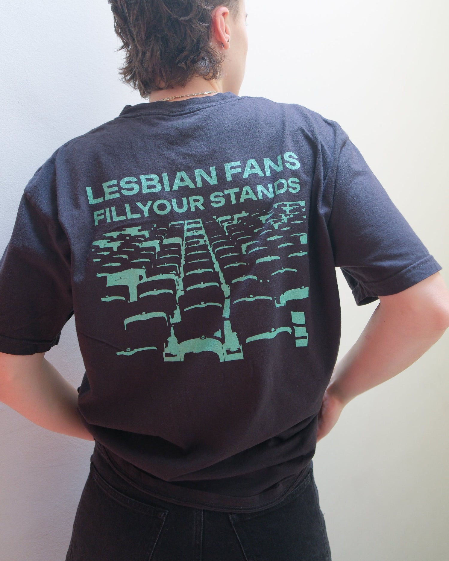 A person with short hair viewed from behind wearing a black graphic t-shirt with the text 'LESBIAN FANS FILL YOUR STANDS' and a green graphic of theater seats, paired with black jeans against a plain light background.